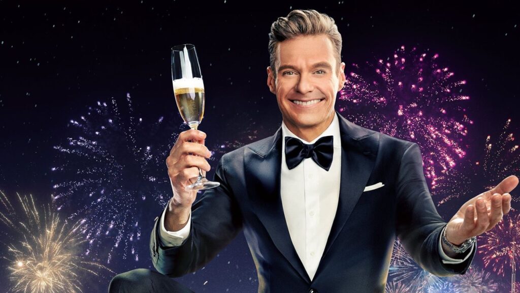 A beaming Ryan Seacrest raises a glass of champagne to welcome in the New Year, dressed in a tuxedo and bow tie and with colorful fireworks exploding in the background.