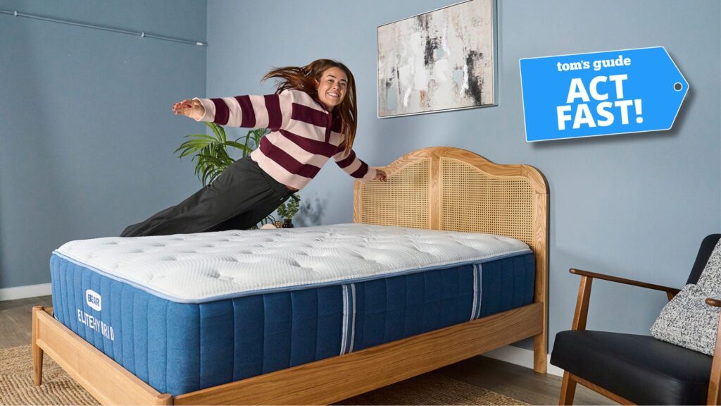 Our mattress tester jumping on the Bear Elite Hybrid mattress on a wooden bedframe with a rattan headboard in our sleep studio with blue walls and a bedside plant with Tom's Guide 'Act Fast' logo