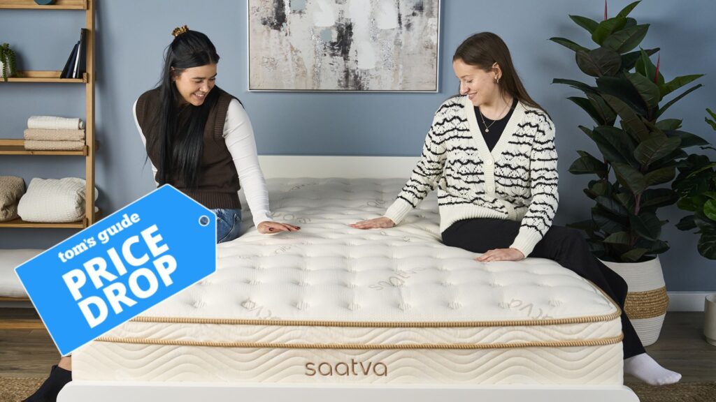The image shows two women sitting on the Saatva Classic mattress, which is placed on a white bed foundation in a blue bedroom