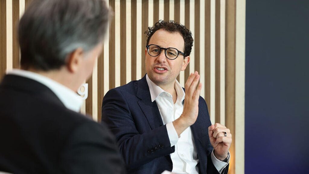 Anthropic CEO Dario Amodei speaks while seated at a conference forum, with the back of another person's head out of focus in the foreground.