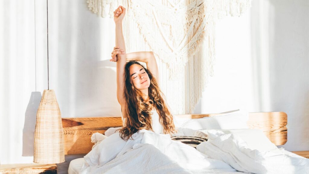 For months I was waking up exhausted — but these 5 easy sleep hacks now help me spring out of bed with bags of energy A woman with long dark hair sits up in bed with her arms stretched in the air as sunlight streams in through her open curtains
