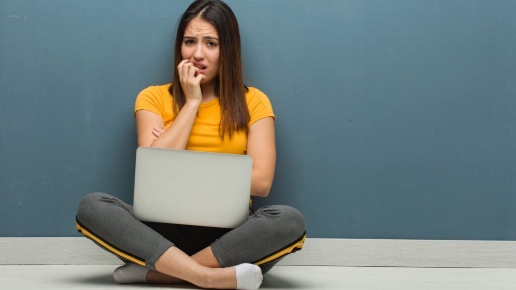 Young woman sitting on the floor with a laptop biting nails, nervous and very anxious