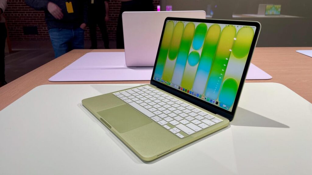 A MacBook Neo in the yellow color named Citrus sits at an angle on a white table. Behind it, the Neo is featured in the pink colored named Blush.