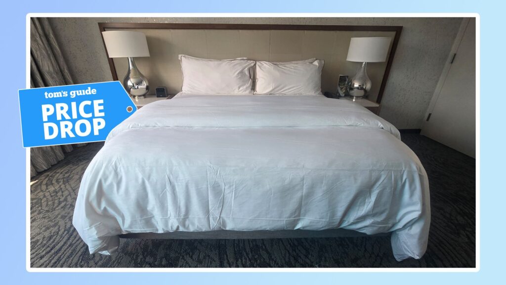 The Westin Heavenly Bed Mattress on a bed frame dressed in white bedding, in a Westin Hotel room. A Tom's Guide price drop graphic sits on the left-hand side and a blue border surrounds the picture