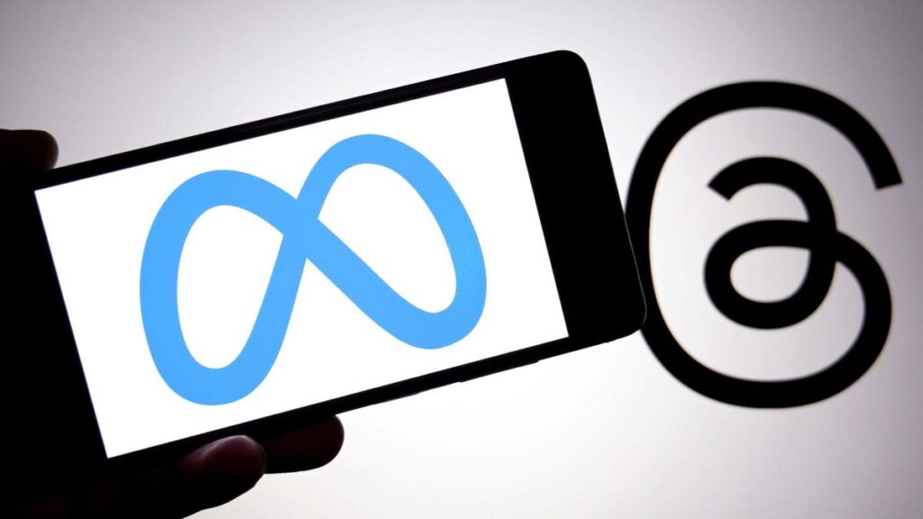 A blue Meta logo on white background on a mobile phone is held next to a black Threads logo.