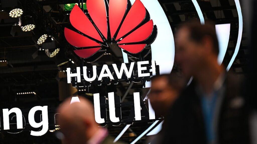 Huawei's logo hangs from the ceiling as several blurry people pass by the camera in the foreground.
