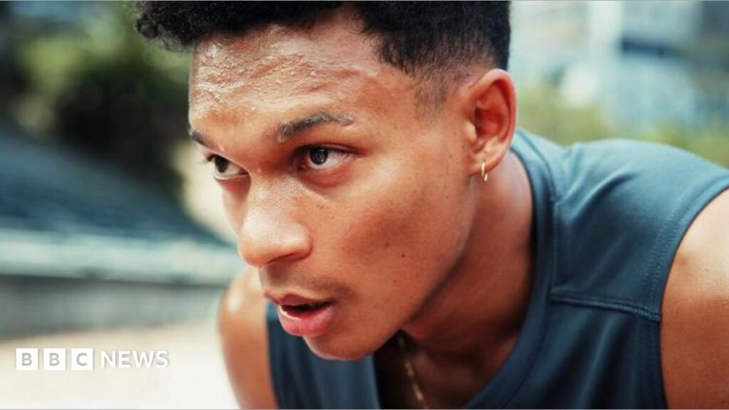 A young man, wearing a sports vest top, is out for a run. He is hunched over, hands on knees, sweating.