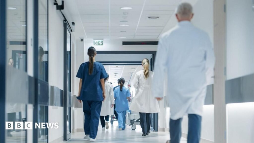 Hospital hallway with doctors, nurses and specialists with their back to camera. They are in a variety of hospital uniforms - dark and light blue scrubs, white coats.