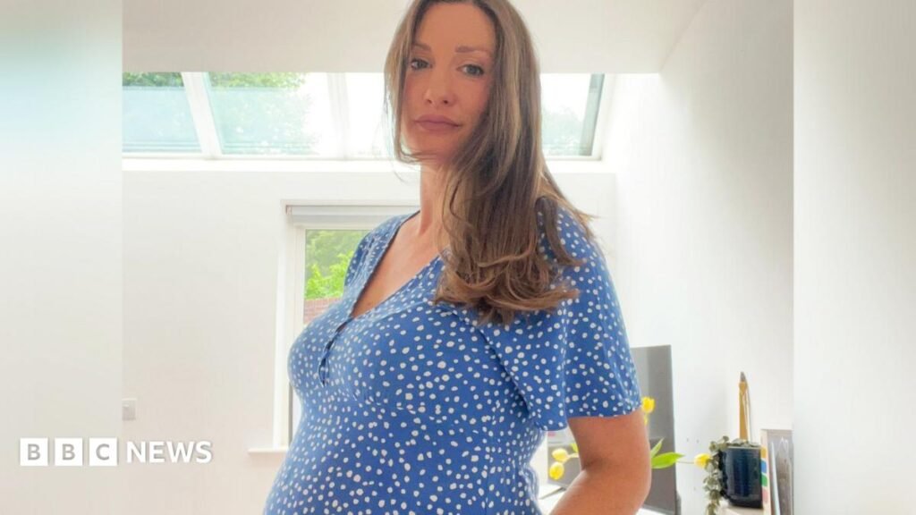 A pregnant woman standing up in a room with white walls and a window behind her. She has long light brown hair and is wearing a blue dress and white dots.