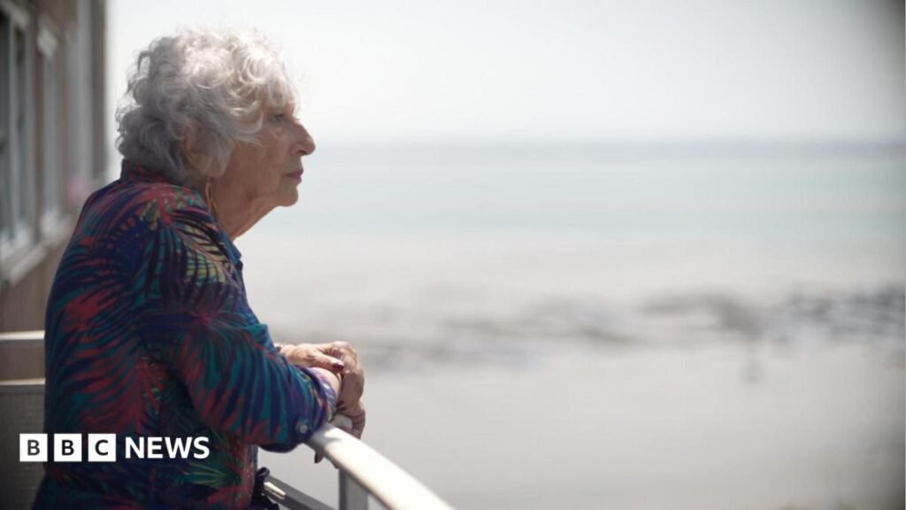 Carol Turansky standing on a balcony. She is wearing a leaf print dress and you can see her side profile. She has grey permed short hair and you can see the sea in the background. She is looking out to the horizon.