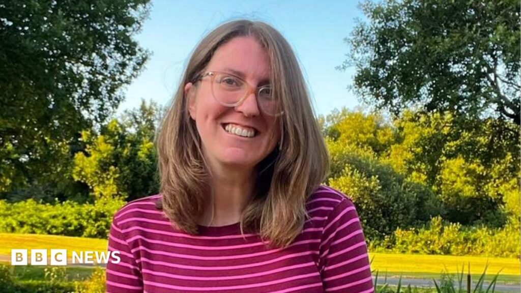 Head and shoulders shot of young woman with glasses and shoulder-length light brown hair. She wears a pink striped top and is surrounded by countryside.