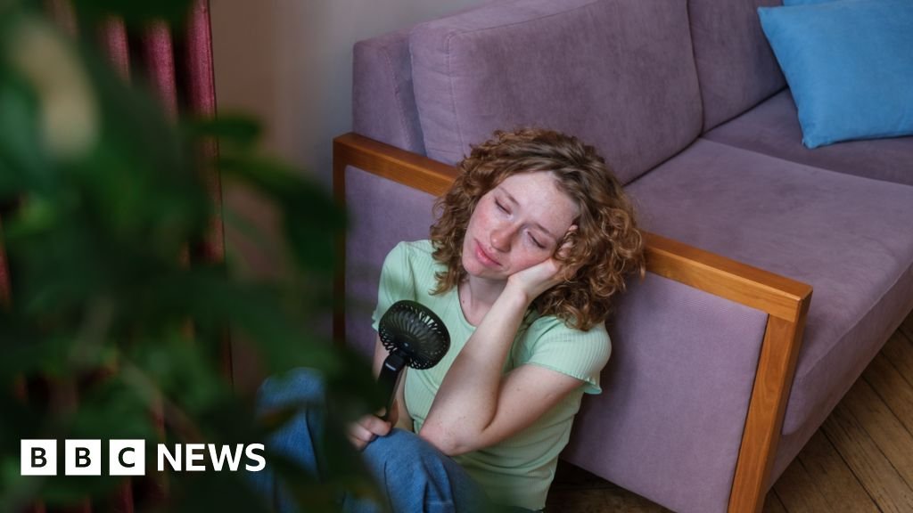 How deadly have the heatwaves in England and Wales been? A woman with shoulder-length curly hair leans on a couch while holding a fan