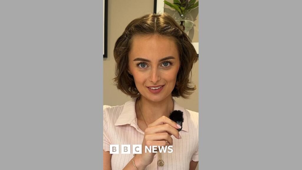 Young woman with short brown hair, smiles at camera, while holding a small clip mic in her right hand. She is wearing a pink short sleeved t-shirt.