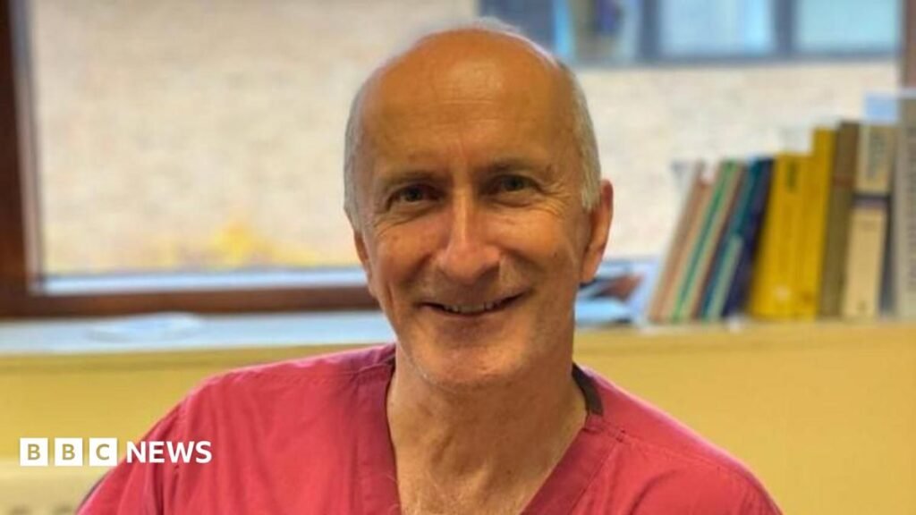 A man in red scrubs smiles into camera, he has white hair on the sides of his head