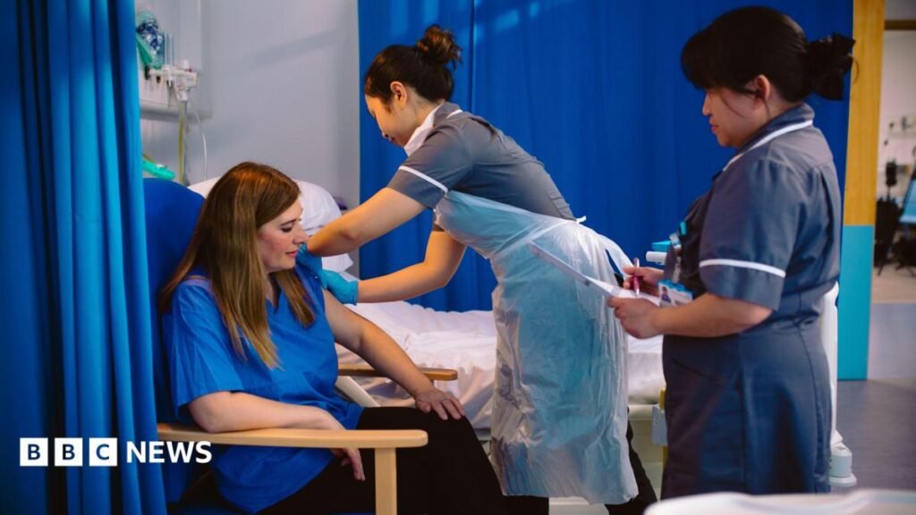 This image shows a healthcare setting with a seated adult receiving an injection in the upper arm from a healthcare worker wearing gloves and a disposable protective apron. A second healthcare worker stands nearby holding paperwork or a clipboard, appearing to document or monitor the procedure. The scene takes place beside a hospital-style bed and is partially enclosed by blue privacy curtains. The color palette is dominated by blue, grey, and white tones, with bright indoor lighting illuminating the room and medical equipment visible in the background. The photograph focuses on the administration of a vaccine and the interaction between healthcare staff and the seated patient.