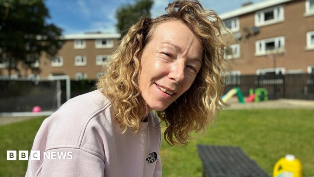 A woman with curly blonde hair smiles outdoors on a sunny day. She’s wearing a pale pink sweatshirt with a visible “The North Face” logo. Behind her, a grassy area features a football goal, colourful playground equipment, and residential buildings with white-framed windows. The bright light and relaxed pose create a friendly, community atmosphere.