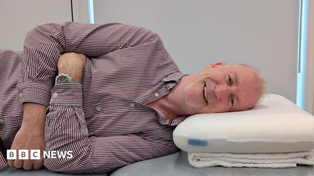 Person lying on their side on a padded treatment table with their head resting on a foam pillow. The person is wearing a checked button-up shirt and a wristwatch, with arms folded across the torso. The pillow is positioned on a folded towel. The window blinds in the background are drawn.