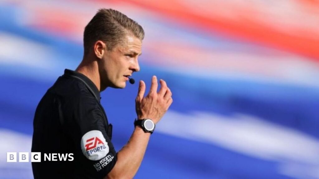 Dan Robathan has short blonde hair and is wearing a referee kit. He has a whistle in his mouth.
