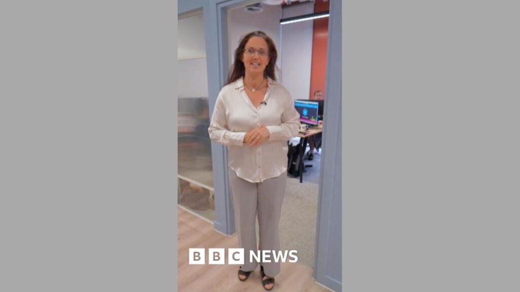 A woman in light clothing with her hands folded standing in the doorway looking at the camera.
