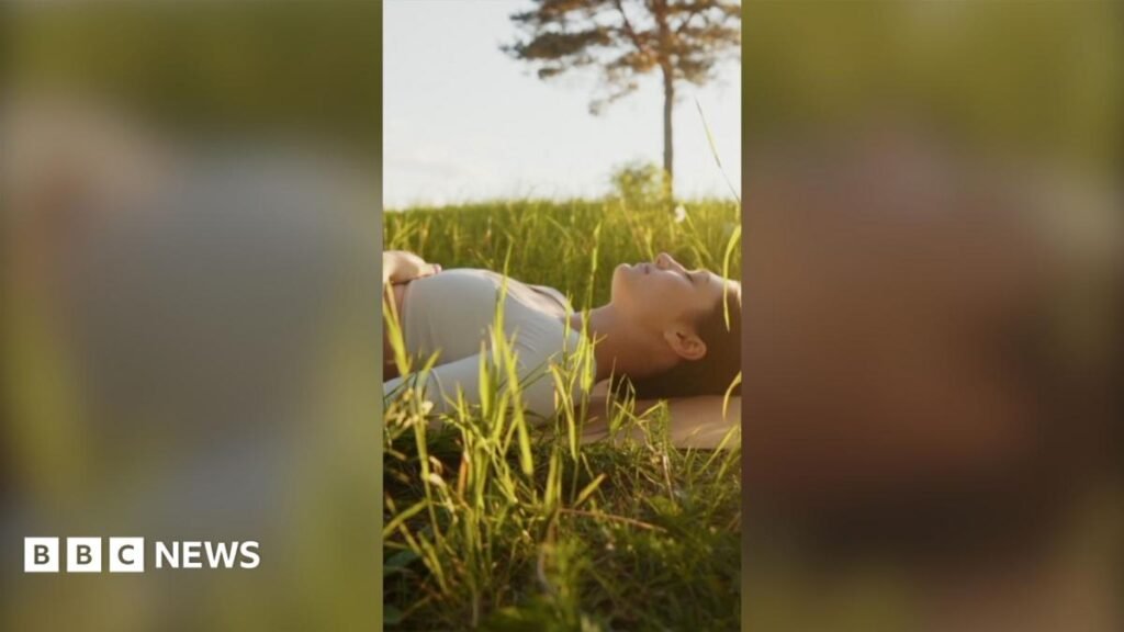 How 'box breathing' can help with stress A woman laying down in the grass outside. It's a sunny day and she looks calm and has her eyes closed. There is a green tree in the background.
