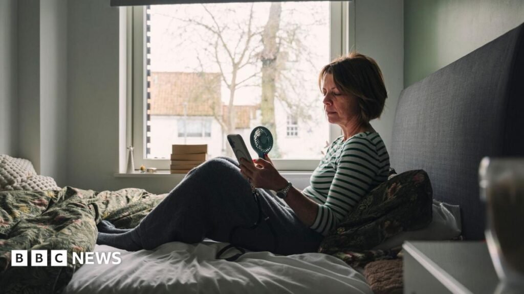 A stock photo of a woman in bed with a handheld electric fan.