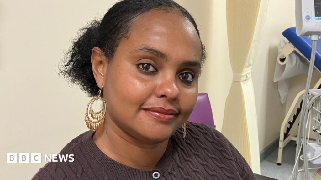 A woman with black hair, tied back, large gold earrings is smiling at the camera.  She is wearing a brown cardigan.