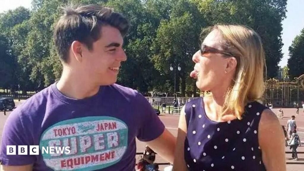Oliver McGowan, wearing a purple t-shirt with a logo, is standing outside Buckingham Palace. He is starring at his mother Paula McGowan who is standing next to him and wearing a dark blue polka dot dress and sunglasses. She is playfully sticking her tongue out at her son and he is laughing.