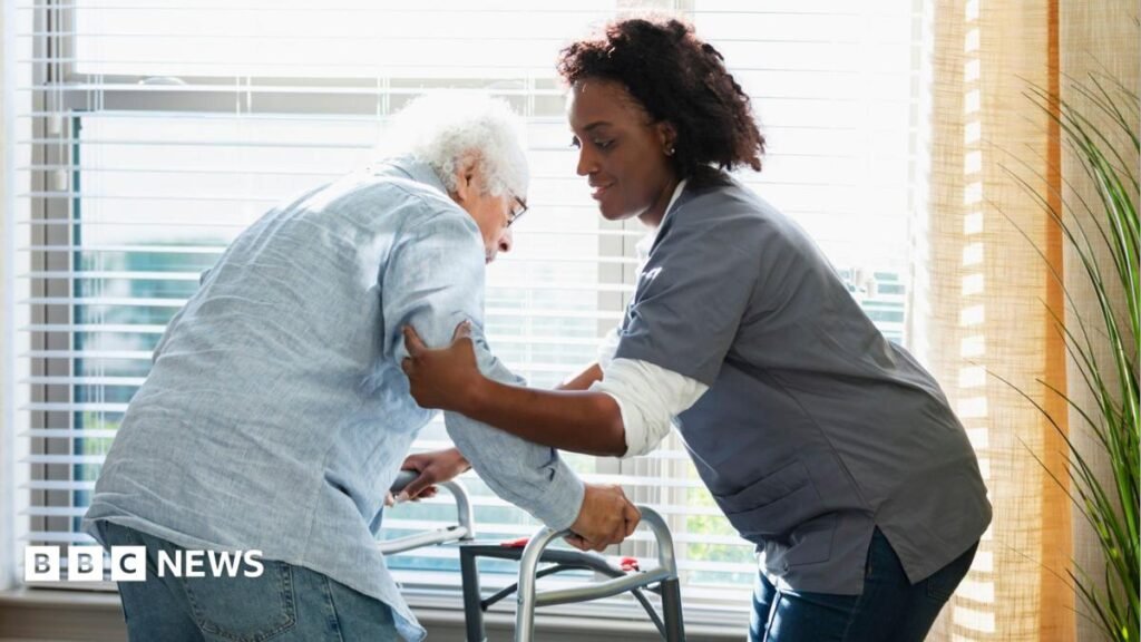 New NHS-style social care system could take decades, Dilnot says A female care home worker, wearing nursing uniform, helps support an elderly man leaning on a walking frame.
