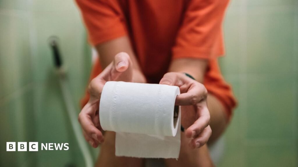 A teenage girl sits on the toilet, holding toilet paper in her hands
