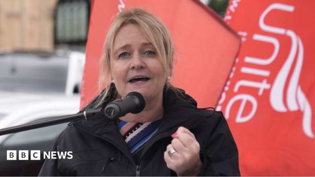 Head and shoulders shot of Graham, who has blonde hair pulled back in a ponytail and is wearing a black raincoat over a striped blue and black top. She has her fist clenched with red nail varnish visible and two wedding rings. She is standing in front of a red Unite the Union flag.