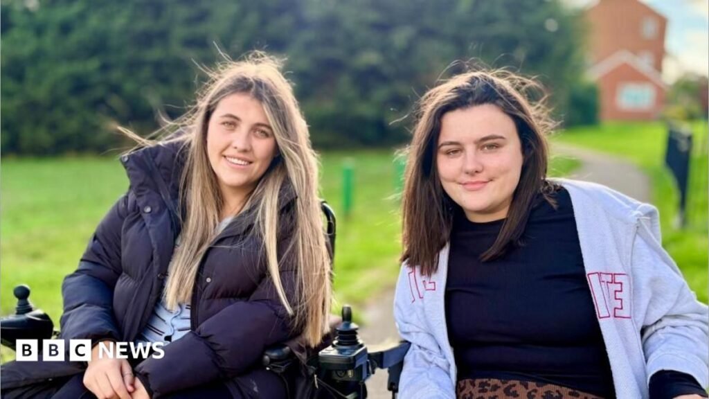 Two women in wheelchairs smile at the camera. There is a path and an area of green grass behind them. The woman on the left has long blond hair and is wearing a pale blue cardigan with a black puffa coat over the top. The woman on the right has shoulder length dark brown hair and is wearing a black top and an open, grey zip-up hoodie.