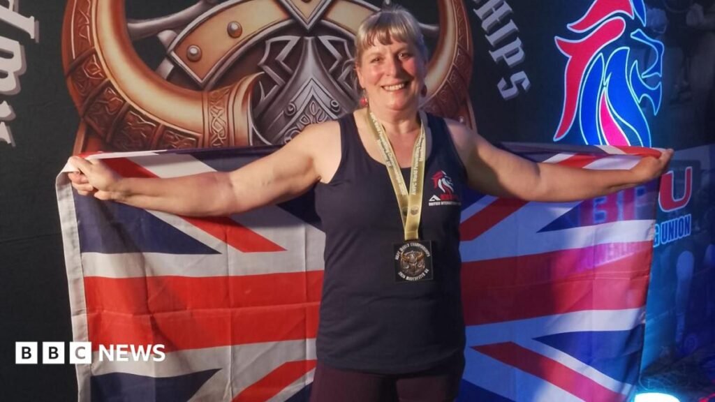 The Deadlifting Grandma: Martine Barons, 65, is triple world champion Martine Barons stood on a podium smiling with both her arms up in the air. A line of women stand behind her looking up to her smiling. Behind is a black board that says 'AWPC World Champion'