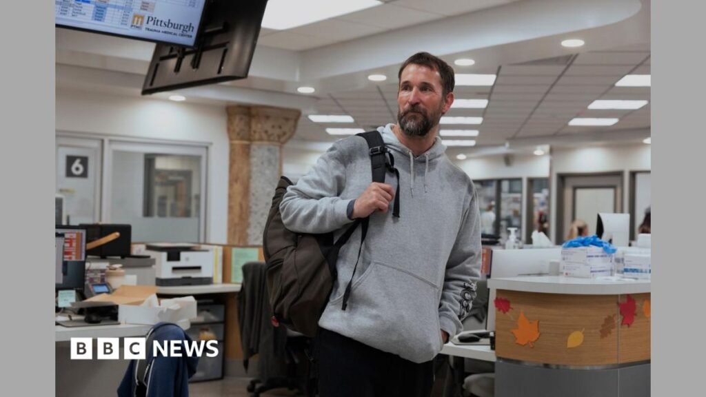 Dr. Michael “Robby” Robinavitch (Noah Wyle) looking exhausted, wearing black scrubs and holding a door open