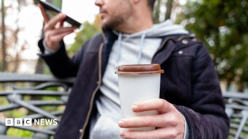 Why caffeine makes some people buzz A close of shot of a man holding a takeaway coffee cup while speaking on his phone. He is wearing a blue jacket and grey hoodie.