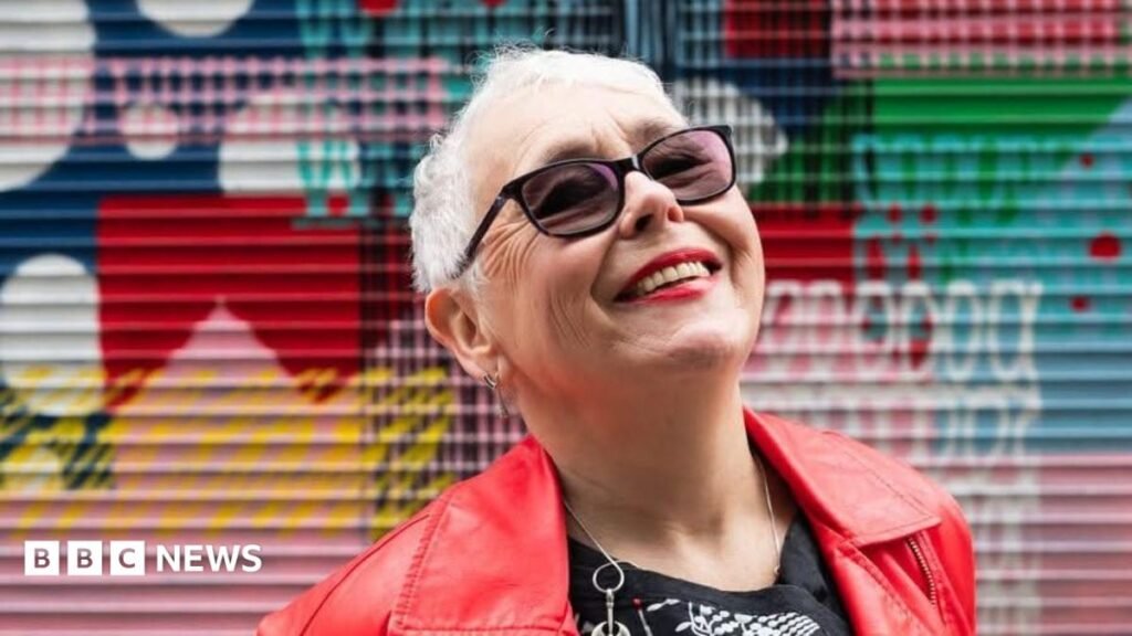 A tightly cropped photo of a woman in her 70s with short grey hair looking up at the sky, smiling. She has sunglasses on, and bright red lipstick that matches her leather jacket. Graffiti is in the background, slightly out of focus.