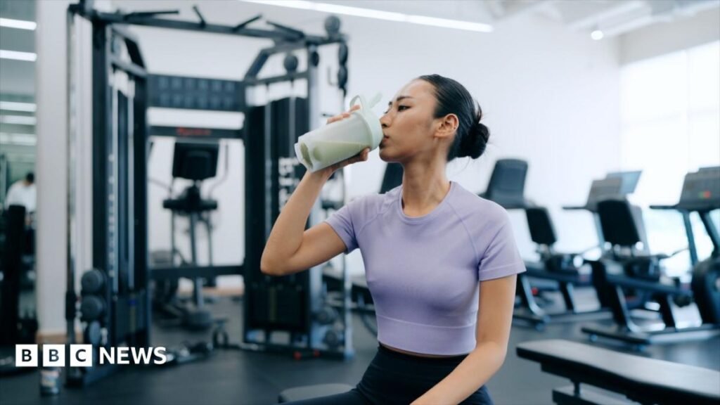 A young woman is sat in a gym drinking a protein shake. She is wearing a purple cropped top and navy blue leggings.