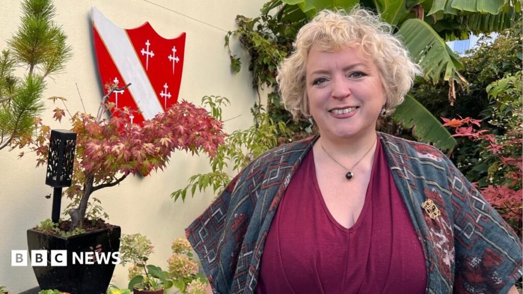 Charlotte Howard in a busy garden smiling at the camera, she has short blond hair and wears a maroon V neck top and blue and red shawl.
