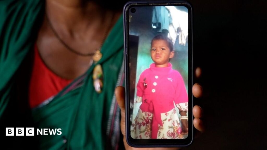A woman in a green saree sits in a dark hut, holding up a smartphone showing the picture of a little girl
