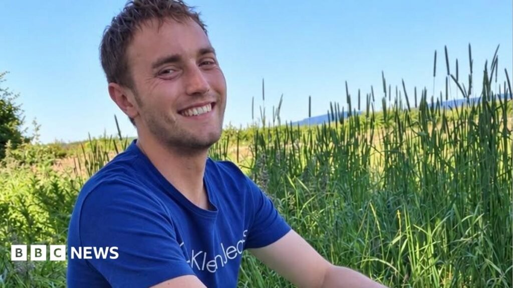 Hugo Flint Cahan is looking into the camera smiling. He is sat in a field surrounded by long grass on a bright summer day. He is wearing a blue t-shirt.